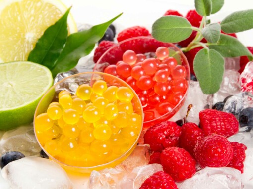 Perles de fruits rafraîchissantes – Une composition colorée mettant en valeur des perles de fruits jaunes et rouges servies sur un lit de glace, entourées de framboises, myrtilles, citrons et feuilles fraîches. Un mélange de saveurs acidulées et sucrées parfait pour des desserts ou des boissons gourmandes.