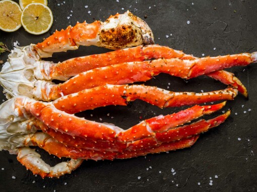 Des pattes de crabe royal rouges et blanches disposées sur une surface sombre, avec des grains de sel et des épices parsemés autour. En arrière-plan, des tranches de citron et une branche d’herbes aromatiques accompagnent le crustacé, prêt à être dégusté.
